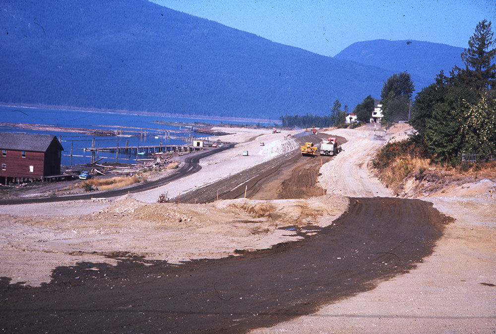 A colour photograph showing land being levelled and cleared beside a waterfront. Construction vehicles and cars in the distance. A building sits by the water on the left. Mountains in the background and trees on the right.