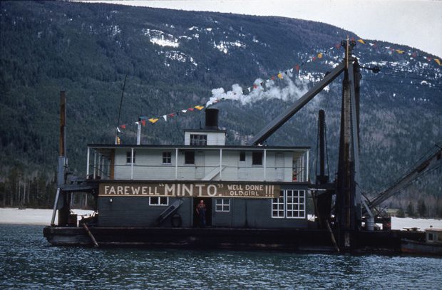 A dredge sits on the water with a sign on the front reading 'Farewell Minto. Well Done Old Girl!!!' A man stands in the doorway under the sign. A banner with colourful triangles is attached to the top of the dredge. A mountain is in the background.