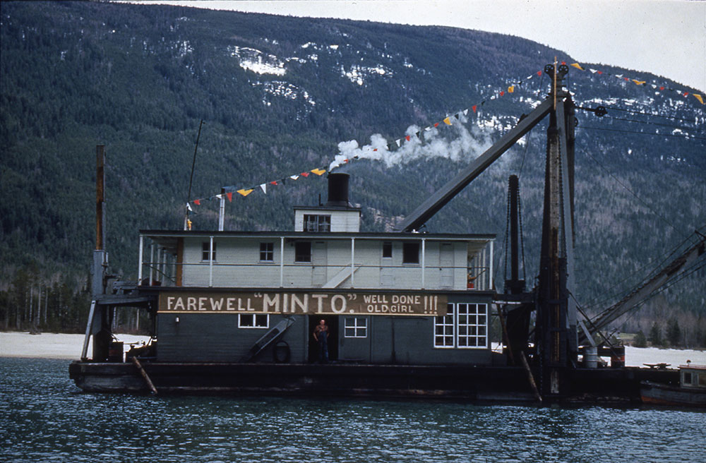 A dredge sits on the water with a sign on the front reading 'Farewell "Minto." Well Done Old Girl!!!' A man stands in the doorway under the sign. A banner with colourful triangles is attached to the top of the dredge. A mountain is in the background.