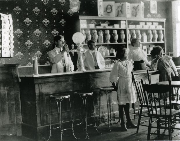 A black-and-white photograph of an ice-cream parlour room. Two men and one women stand behind a bar-counter with an ice-cream machine on top, and bar stools in front. Two girl stand in front of the counter behind a table and chairs. The wall is covered in wallpaper, and has a shelf full of items in front of it.