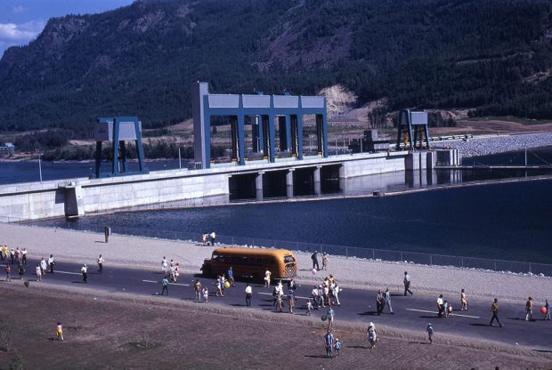 A crowd of people and a bus are on the road in front of the dam. Part of a mountain is in the background.