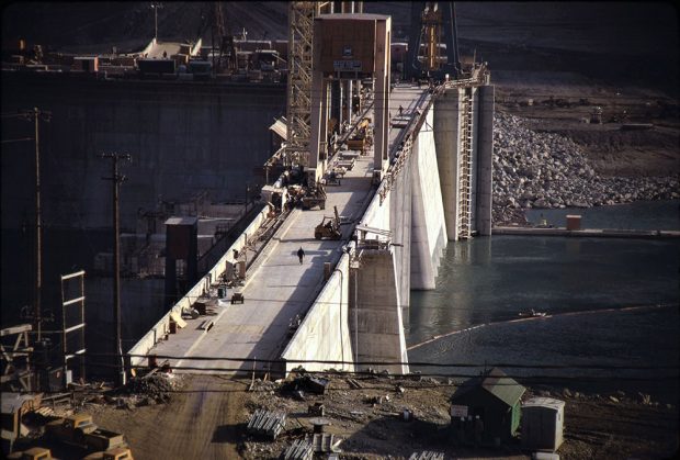 A close-up photograph of the dam under construction, nearly complete. People and construction vehicles are on the dam.