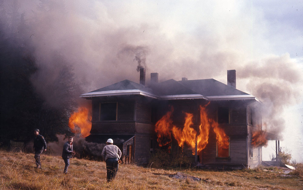 A colour photograph showing three people standing in front of a burning house. The house is still intact with orange flames coming out of the windows. Thick white and black smoke surrounds the house.