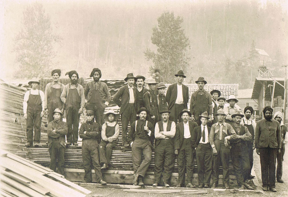 A grayscale photograph of a group of men, a work-crew, standing in front of a pile of wood. The men were from Japan, India, and across Canada and Europe. Trees in the background of the photo..