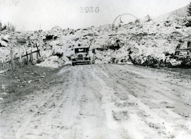 A black-and-white photograph of a avalanche. The snow is piled up in the background. A car sits in front on the road. There is a fence on either side of the road.