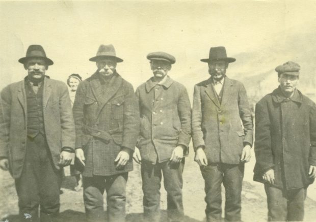 A grayscale photograph of five men standing in a row. The men are wearing jackets and hats, and all have facial hair. A woman can be seen being on the left.