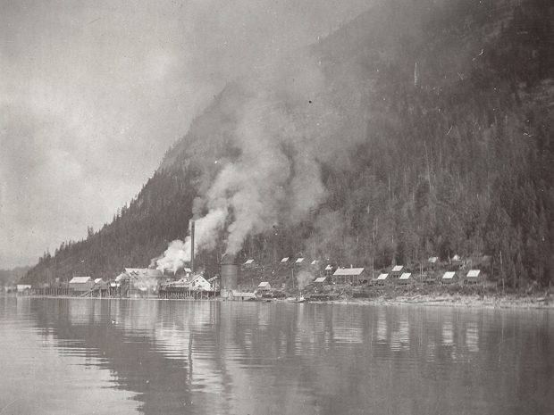 A black-and-white photograph of a sawmill sitting behind a body of water and in front of a mountain. Smoke is coming out of the mill. There are houses beside the mill.