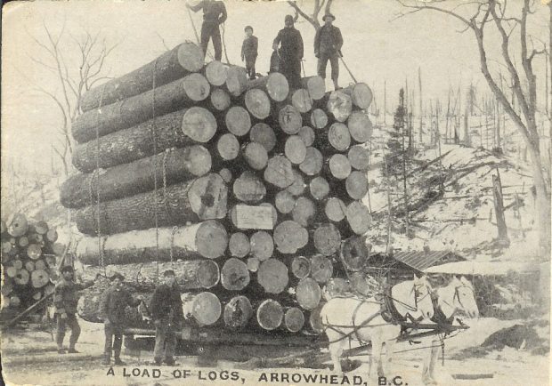 Four people stand on top of a tall stack of logs sitting on a crate attached to two horses. Three men stand to the left of the logs. A tree-clearing is in the background. The bottom of the photograph has text A load of logs, Arrowhead, B.C.