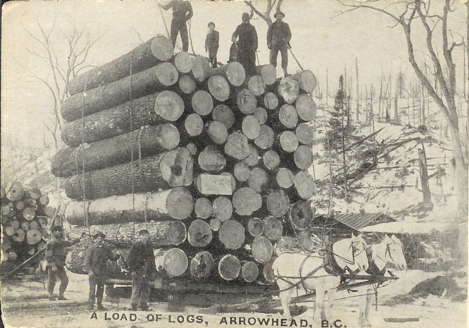 Four people stand on top of a tall stack of logs sitting on a crate attached to two horses. Three men stand to the left of the logs. A tree-clearing is in the background. The bottom of the photograph has text "A load of logs, Arrowhead, B.C."