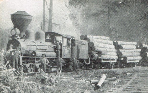 A black-and-white photograph of a train hauling loads of logs. Six men stand around the front of the train, and the conductor is sticking his head out of the window. Trees in the background.