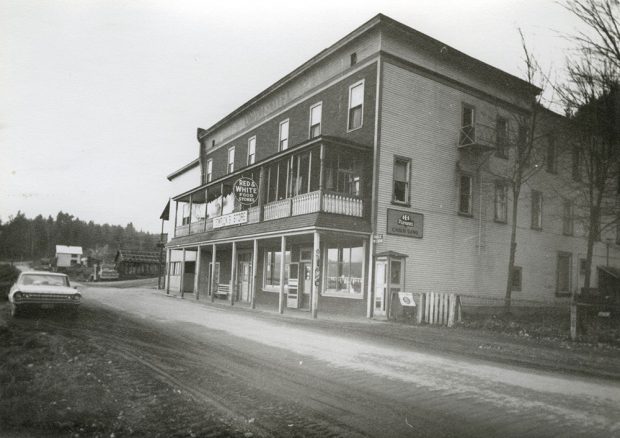 A black-and-white photograph of a two-story building next to a dirt road. The building has lettering at the top that reads Union Hotel and a couple signs at front, one that read Red & White food store and the other Temricks Store. A car sits on the opposite side of the road. Buildings and trees in the distance.