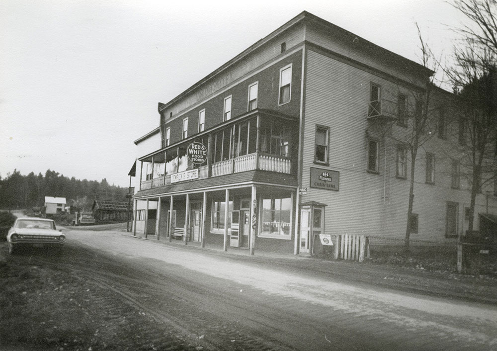 A black-and-white photograph of a two-story building next to a dirt road. The building has lettering at the top that reads "Union Hotel" and a couple signs at front, one that read "Red & White food store" and the other "Temricks Store". A car sits on the opposite side of the road. Buildings and trees in the distance.