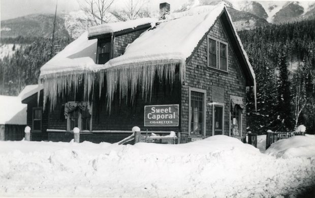 A black-and-white photograph of a building in a winter scene. Long icicles hang from the side of the building. A sign reading Sweet Caporal Cigarettes is to the left of the building. The store is surrounded by snow. Snowy trees and the base of a mountain are in the background.