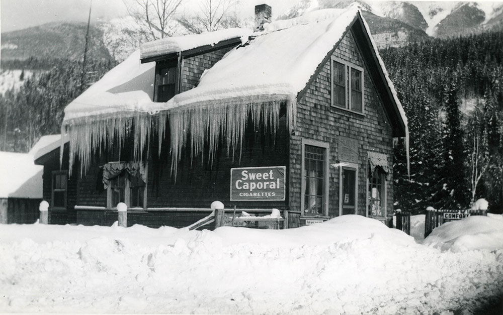 A black-and-white photograph of a building in a winter scene. Long icicles hang from the side of the building. A sign reading "Sweet Caporal Cigarettes" is to the left of the building. The store is surrounded by snow. Snowy trees and the base of a mountain are in the background.