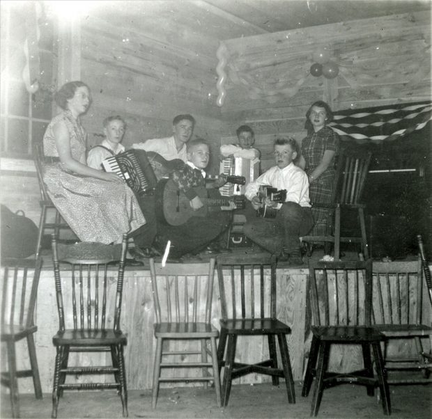 A black-and-white photograph of young people sitting on a stage holding instruments. Three boys are holding guitars, and two are holding accordions. Two ladies on either side. There are streamers and balloons on the wall behind the stage, and a row of chairs in front of the stage facing outwards.