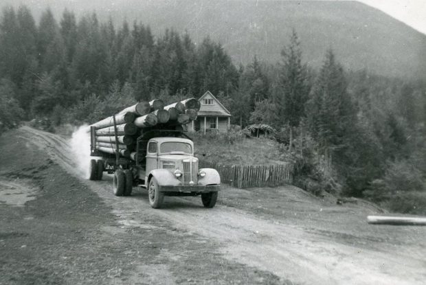 A black-and-white photograph of a truck hauling log poles down a dirt road. A house is behind the truck. Trees and the base of a mountain in the background.