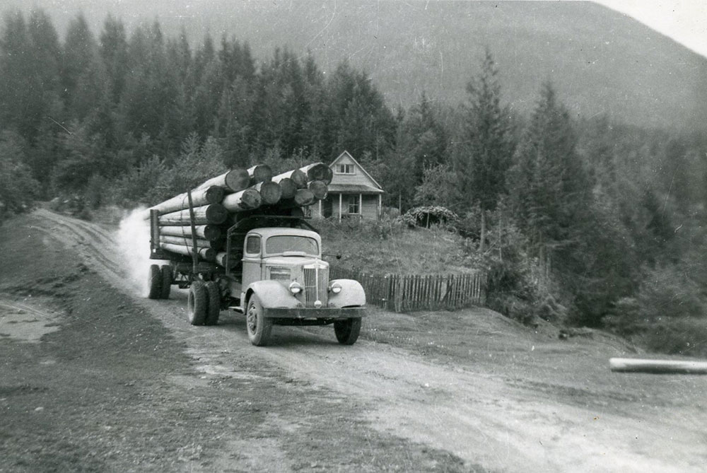 A black-and-white photograph of a truck hauling log poles down a dirt road. A house is behind the truck. Trees and the base of a mountain in the background.