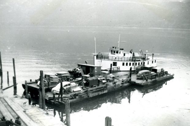 A black-and-white photograph of a barge with cargo, including four vehicles. The barge sits on the water, and a part of the dock can be seen on the left. Four people and two dogs stand on the dock.