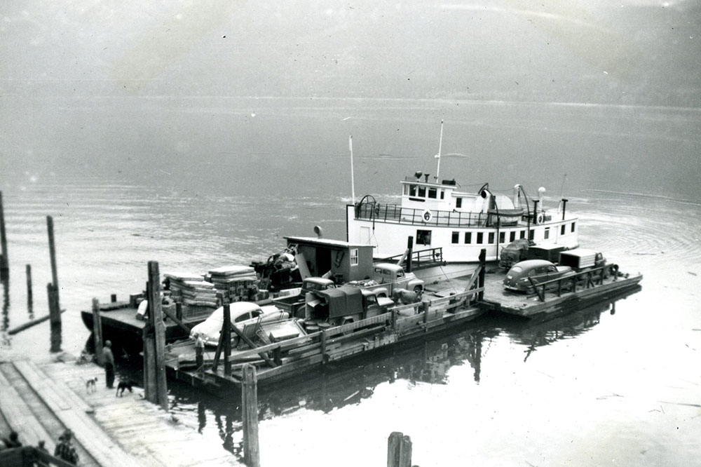 A black-and-white photograph of a barge with cargo, including four vehicles. The barge sits on the water, and a part of the dock can be seen on the left. Four people and two dogs stand on the dock.