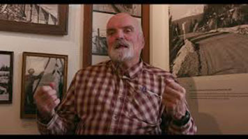 Bill Green – a bald white man with short grey beard. Wearing a red plaid shirt. He is sitting in a chair in front of the corner of a room. There are black and white photos on the wall.