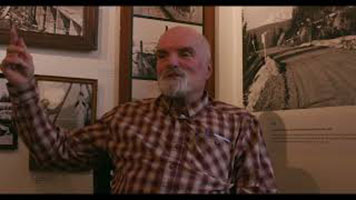 Bill Green – a bald white man with short grey beard. Wearing a red plaid shirt. He is sitting in a chair in front of the corner of a room. There are black and white photos on the wall.