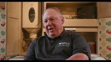 Walter Bobicki - a white man with grey hair and a black collared, short sleeve shirt. He is sitting in the Stories Beneath the Surface exhibit room in front of the kitchen set. There is cherry wallpaper behind him.