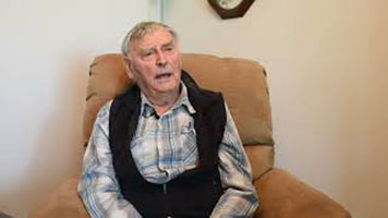 Harold Catherwood. Harold Catherwood is an elderly white man with short grey hair. He is wearing a printed collared shirt and a black vest. He is sitting on a light brown armchair in a room with black and white clock on a white wall.