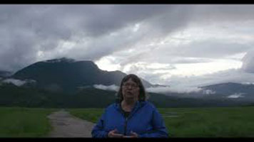 Cathy English - a white woman with short grey hair, wearing a blue zip-up sweater. She is standing on a dirt path. There is green grass, a mountain, and grey sky behind her.