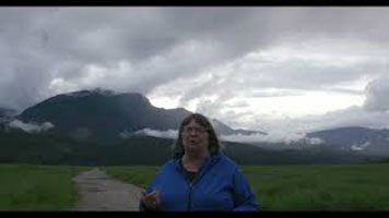 Cathy English - a white woman with short grey hair, wearing a blue zip-up sweater. She is standing on a dirt path. There is green grass, a mountain, and grey sky behind her.