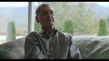 Brian Gawiuk - a white man with grey hair, glasses, and a plaid button down shirt. He is sitting in a living room on a couch, with clear windows behind him and a view of trees in the background.