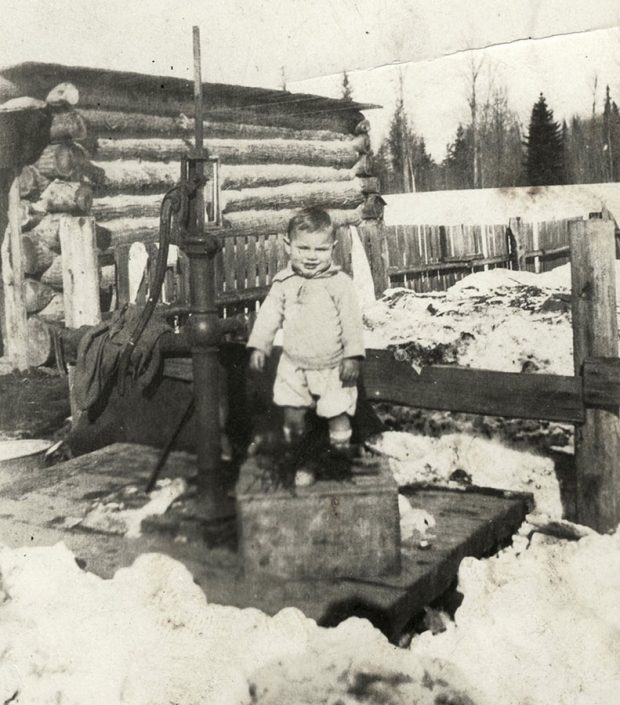 A black-and-white photograph of a small boy standing on top of a crate beside a water pump. There is a fence behind him, and a log building to his left. It is a winter day, with piles of dirty snow on the ground.