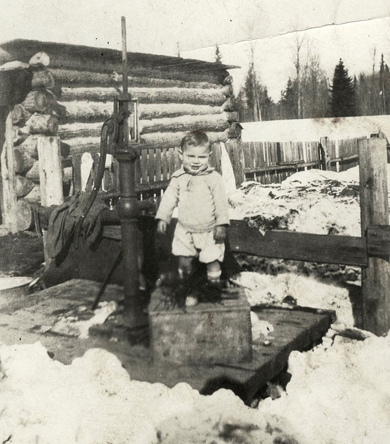 A black-and-white photograph of a small boy standing on top of a crate beside a water pump. There is a fence behind him, and a log building to his left. It is a winter day, with piles of dirty snow on the ground.