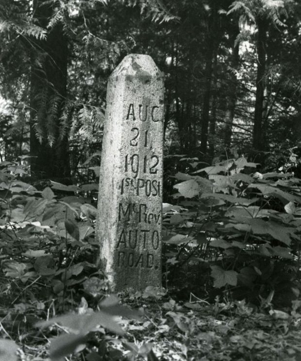 A black-and-white photograph of a tall prism-shaped post surrounded by trees and other greenery. The post reads Aug 21 1912 1st Post Mt. Rev. Auto Road.