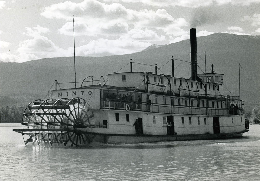 Black-and-white photograph of a steamship, angled left, on the water. The photograph shows a clear-view of the paddle-wheels on the front of the steamship, and the name “MINTO” above it. Mountains and a partly-clouded sky are in the background.