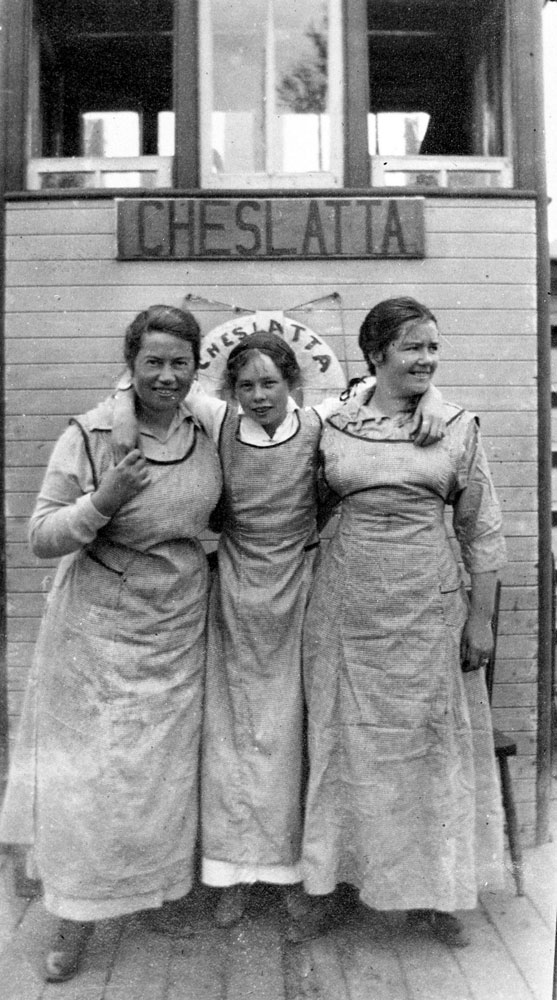 A black-and-white photograph of three women standing together. The woman in the middle has her arms over the shoulders of the other women. They stand in front of a buoy and a sign that both read 'Cheslatta'.