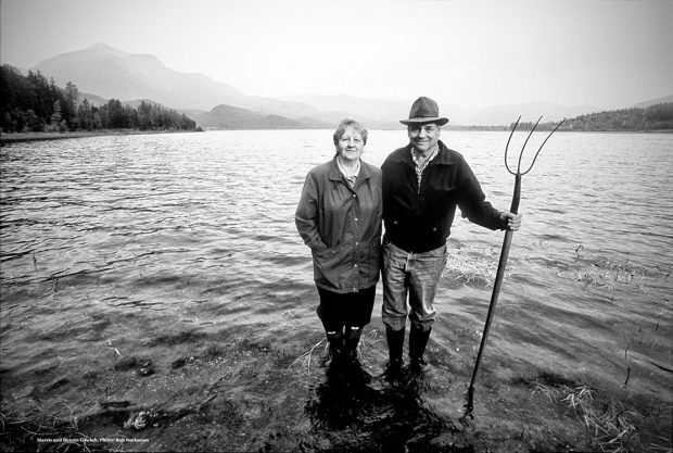 A black-and-white photograph of a woman and a man standing ankle deep in a body of water. The man is holding a pitchfork. Mountains and trees are in the background.