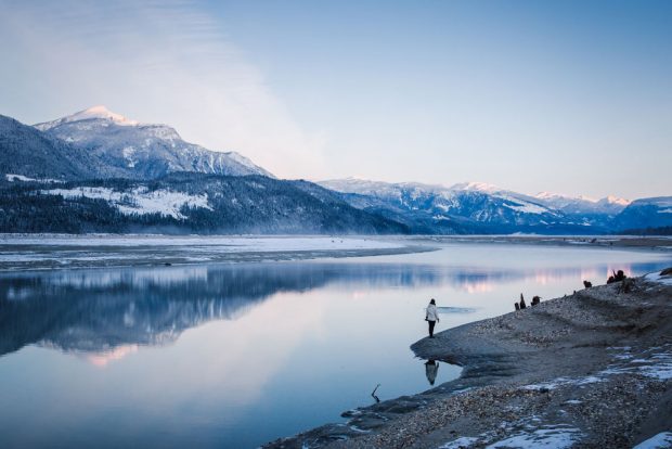 A colour photograph of snowy mountains reflecting off a body of water. A person stands on the bank beside the water.