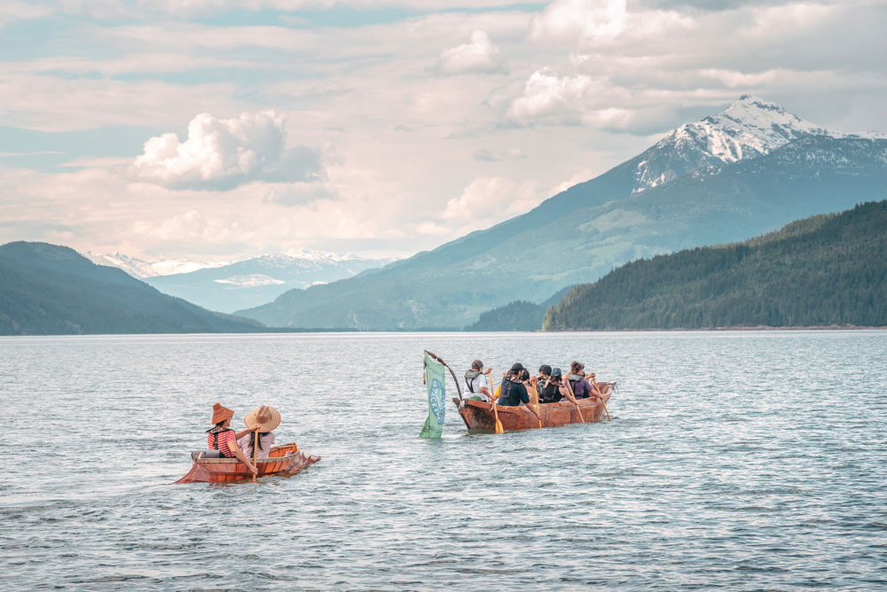 A coloured photograph of two people in a sturgeon-nosed canoe paddling behind a larger dugout canoe, flying the Sinixt flag. The boats are on a body of water with mountains in the background, and a partly-cloudy sky above.