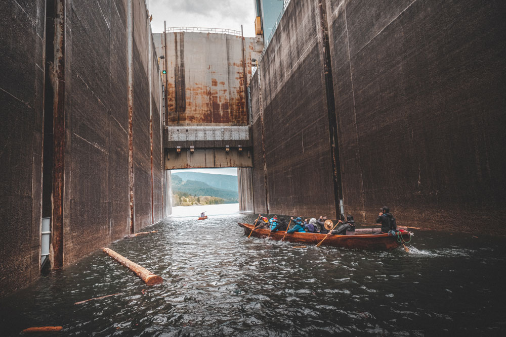 A colour photograph of a group of people rowing a canoe through the dam locks. Some logs sit in the water to the left of the canoe. Mountains can be seen through the open end of the lock gates.