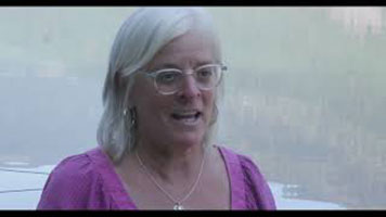 Eileen Pearkes - a white woman with white hair. She is wearing white glasses, a silver necklace, and a pink shirt. She is sitting in front of a body of water.