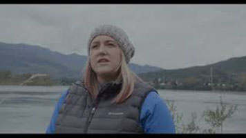 Kate Borucz - a white woman with blond and pink hair. She is wearing a grey toque, a blue long sleeve, and a black vest. She is standing in front of a body of water with mountains behind.