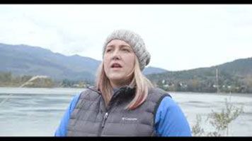 Kate Borucz - a white woman with blond and pink hair. She is wearing a grey toque, a blue long sleeve, and a black vest. She is standing in front of a body of water with mountains behind.