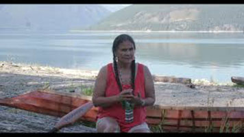 Shelly Boyd – a Sinixt women with dark hair in two braids, wearing an orange tank-top. She is sitting on a log in front of a river.