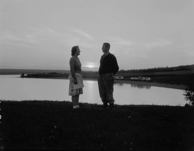 Black and white photograph of a couple standing, facing each other, in front of a lake, at sunset.