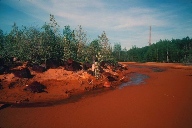 Coloured picture of a polluted body of water, with an orange shade : rusted metal barrels sitting in the forest. In the background, we can see the very tall chimney of a foundry.