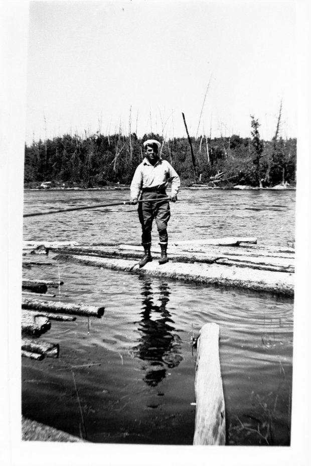 Black and white picture of a log driver on a log afloat.