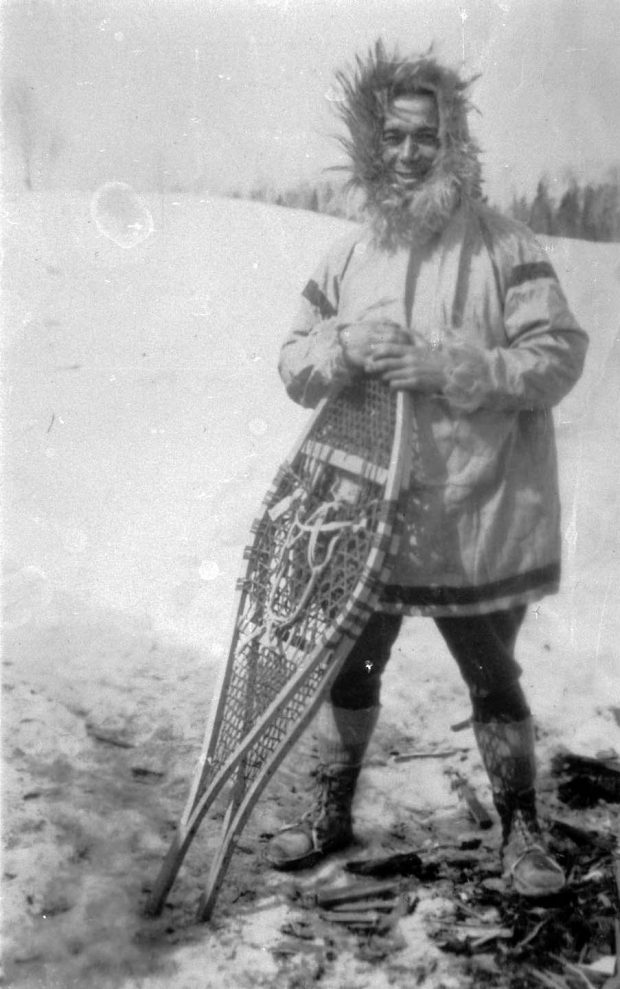 Black and white picture of a man in a parka, snowshoes in hand, outside during the winter.