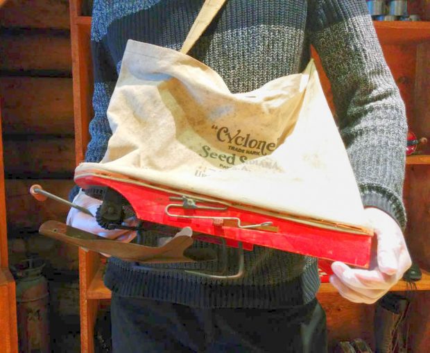 A museum interpreter is holding a red wooden seed planter with a metal rotary distributor and a cotton pouch.