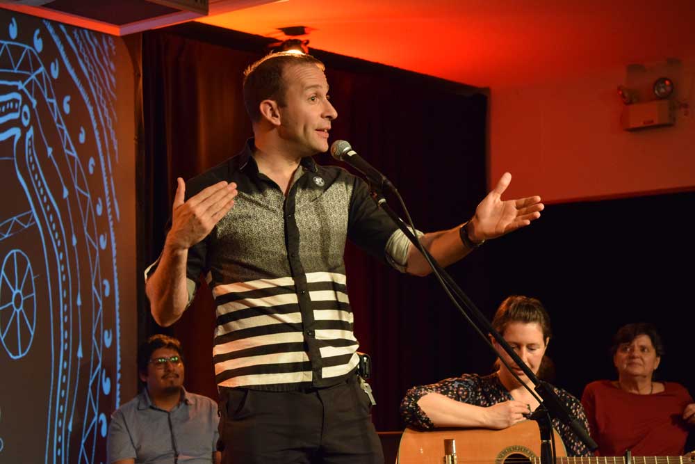 Picture of a man standing on a stage, in front of a mike, telling a story. In the background, two spectators and a woman holding a guitar.