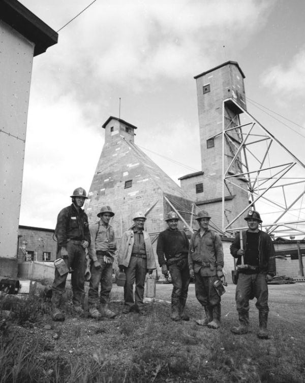 Six miners in work clothes are standing in front of a mine's scaffold and other industrial buildings.